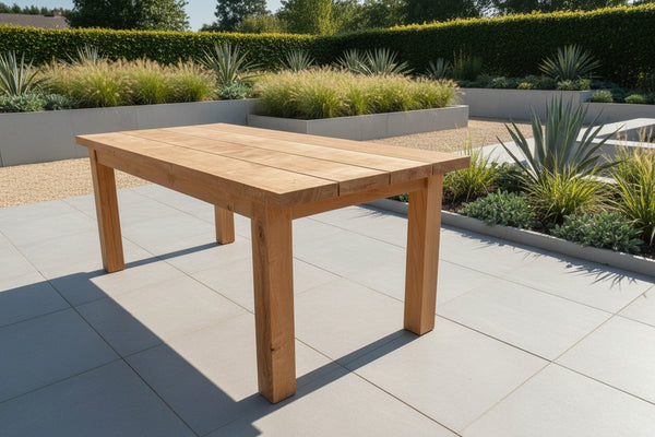 A handmade 4 plank oak garden table with a natural wood finish, displayed on a patio with a manicured landscape in the background.