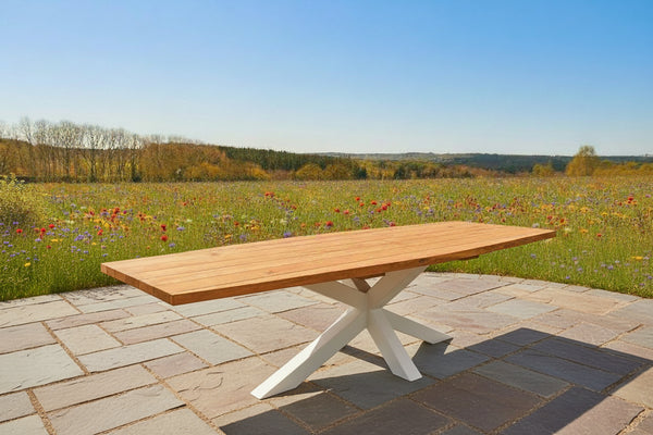 An outdoor dining table with a curved oak tabletop and a white cross-legged steel base, set in an outdoor setting with a cloudy sky in the background.