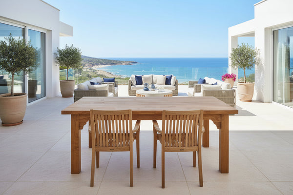A rectangular boarded oak garden table with four square legs on a wooden deck with a sea view in the background.