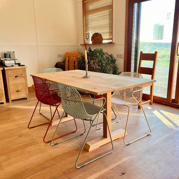 Dining area with a handmade Sussex oak fine pillar dining table by Country Ways and colorful chairs in a room with large windows.