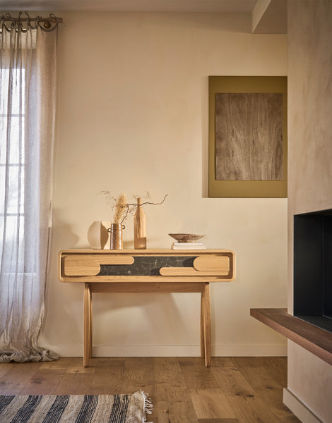 A light wood console table with a dark ceramic front panel and curved drawer pulls, styled in a warm, minimalist room with beige walls, a window with sheer curtains, and a framed abstract print.