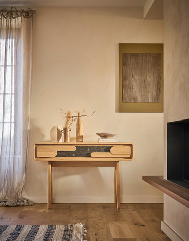 A light wood console table with a dark ceramic front panel and curved drawer pulls, styled in a warm, minimalist room with beige walls, a window with sheer curtains, and a framed abstract print.