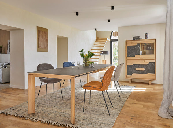 A modern dining room scene featuring a rectangular wooden table with a ceramic top, surrounded by contemporary chairs, set on a textured area rug. The room has light wood floors, off-white walls, a staircase in the background, and a mid-century modern cabinet on the right.