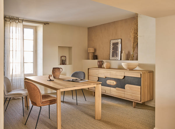 A styled dining room corner featuring a light wood dining table and chairs next to a matching mid-century modern sideboard with dark gray panel accents, set against warm beige walls and textured wallpaper.