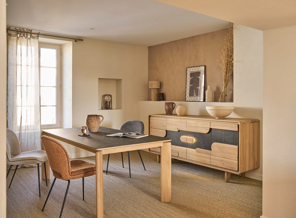 A styled dining room interior featuring a light wood dining table with a ceramic tabletop, matching chairs, a mid-century modern sideboard with wood and dark gray panels, and neutral beige walls and carpet.