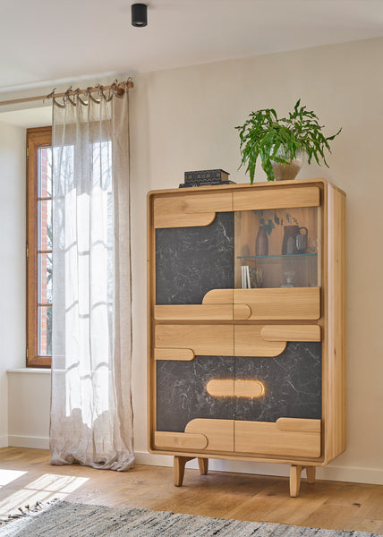 A mid-century modern wooden cabinet with dark ceramic-patterned doors stands in a bright room next to a window with sheer curtains and a potted plant on top.