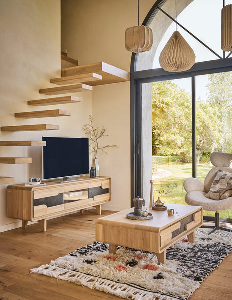 A modern living room featuring a light oak wood TV console, matching coffee table, and a black flat-screen TV mounted on the wall above the console. The room has a floating staircase, large arched windows looking out to a green garden, and natural light.