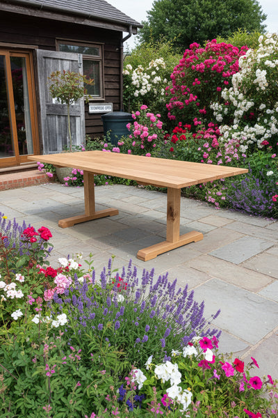 A rectangular wooden dining table with a natural oak finish and T-style legs, set outdoors on a gravel surface in front of a rustic dark wood building.