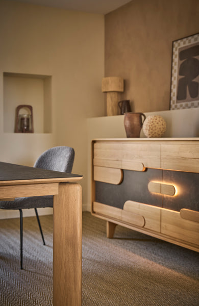 A modern wooden sideboard or credenza in a warm beige and brown tone, styled in a dining room next to a matching wooden table and a gray upholstered chair.
